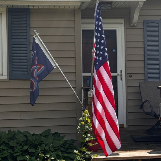 Super Tough 3ft x 5ft Nylon American Flag - Secondary Image - United States Flag Store An American flag mounted on a pole outside a house, with a second dark blue flag hanging to the left, in front of a beige house with shutters and a porch.
