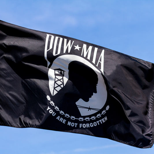 POW/MIA Flag 3x5ft Nylon - Secondary Image - United States Flag Store A black POW-MIA flag with a white printed emblem and the words 'POW-MIA' and 'YOU ARE NOT FORGOTTEN' waving against a blue sky.