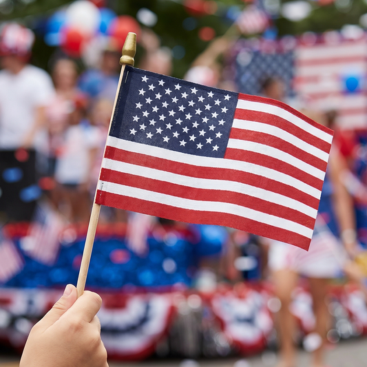 Super Tough 8 A hand holding a small American flag on a wooden stick, with a blurred parade or crowd background in red, white, and blue patriotic decor.