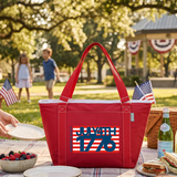 Red cooler bag with 'July 4th 1776' design on a picnic table with people and flags in the background.