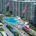 Aerial view of a large resort complex with tall modern buildings, multiple swimming pools, lounge areas, palm trees, and large American flags hanging from the buildings.