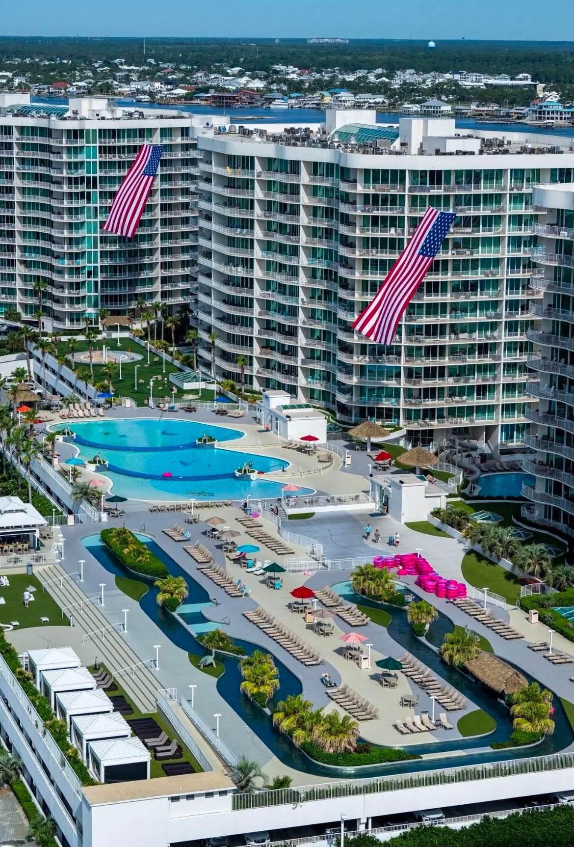 Aerial view of a large resort complex with tall modern buildings, multiple swimming pools, lounge areas, palm trees, and large American flags hanging from the buildings.