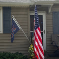 An American flag mounted on a pole outside a house, with a second dark blue flag hanging to the left, in front of a beige house with shutters and a porch.