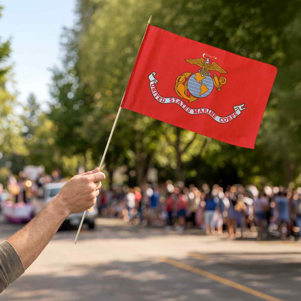 Hand waving a small red United States Marine Corps stick flag at an outdoor parade.