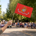 Hand waving a small red United States Marine Corps stick flag at an outdoor parade.