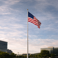 American flag on a flagpole against a blue sky with clouds, buildings, and trees in the background.
