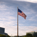 American flag on a pole against a blue sky with clouds, buildings, and trees in the background.