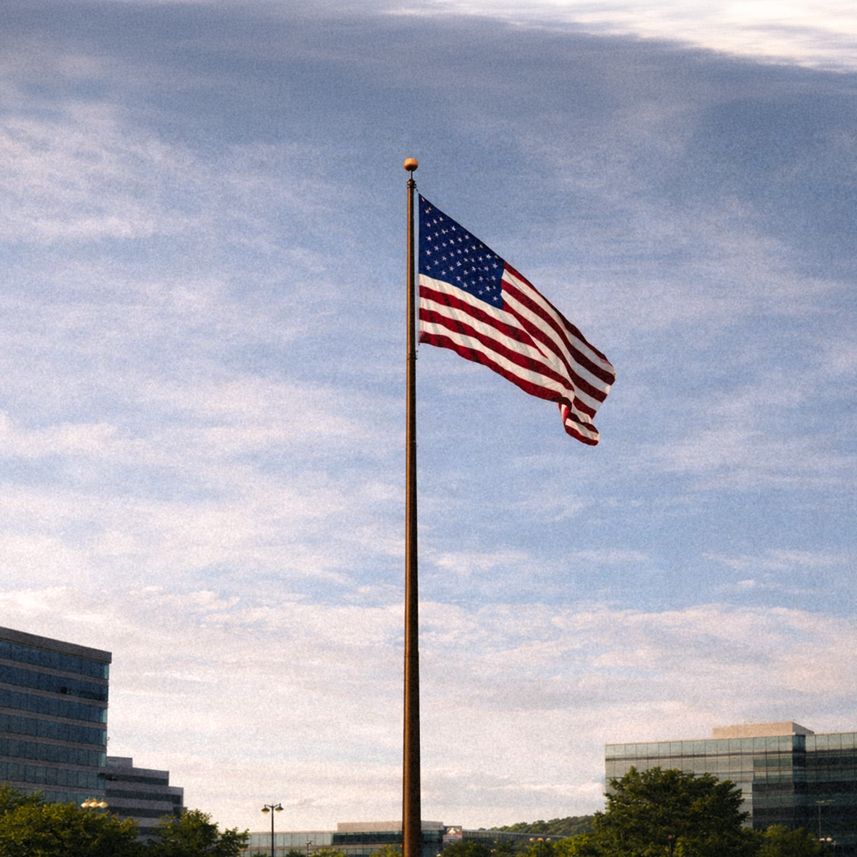 American flag on a pole against a blue sky with clouds, buildings, and trees in the background.