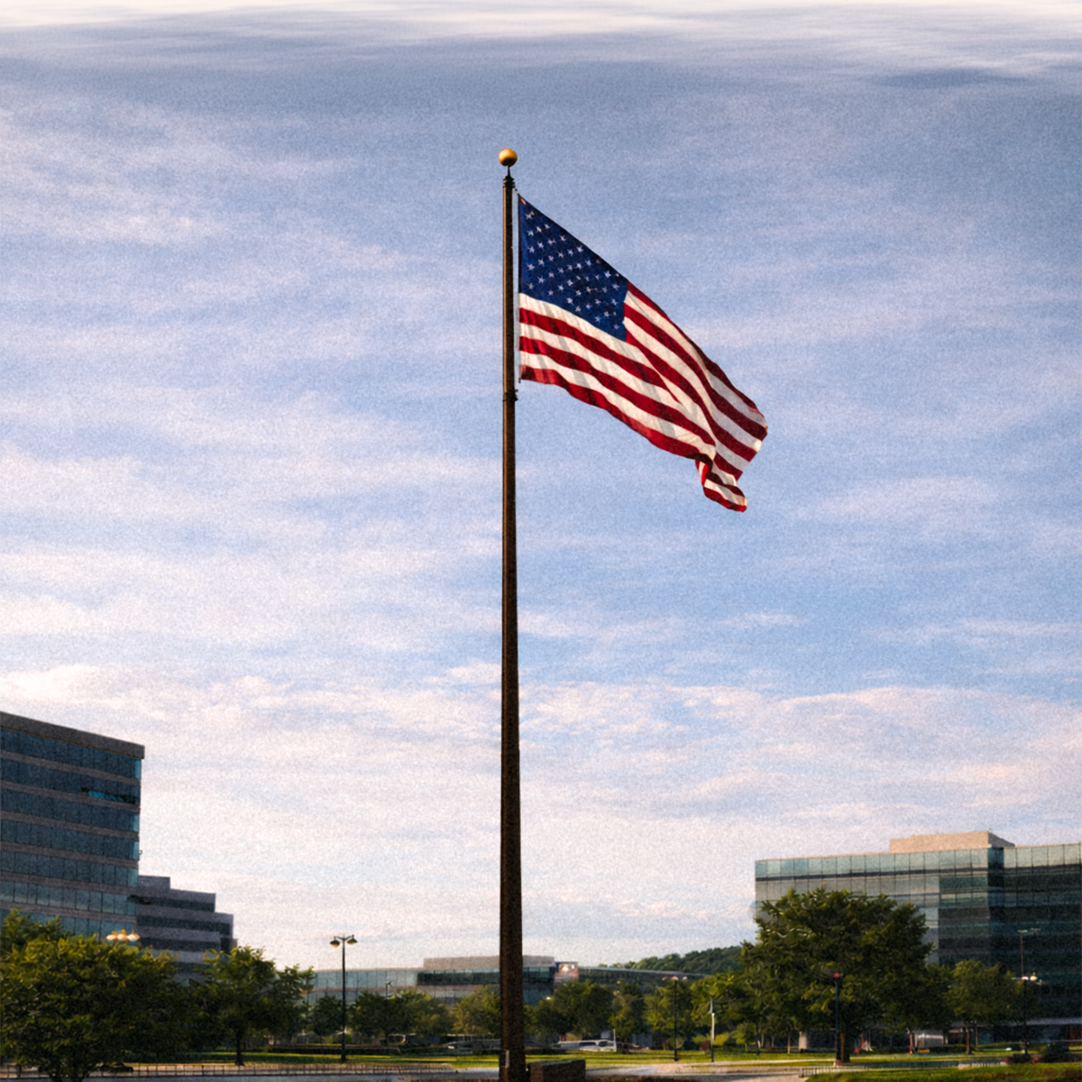 American flag on a flagpole against a blue sky with clouds, with buildings and trees in the background.