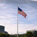 American flag waving on a flagpole against a blue sky with clouds, buildings, and trees in the background.
