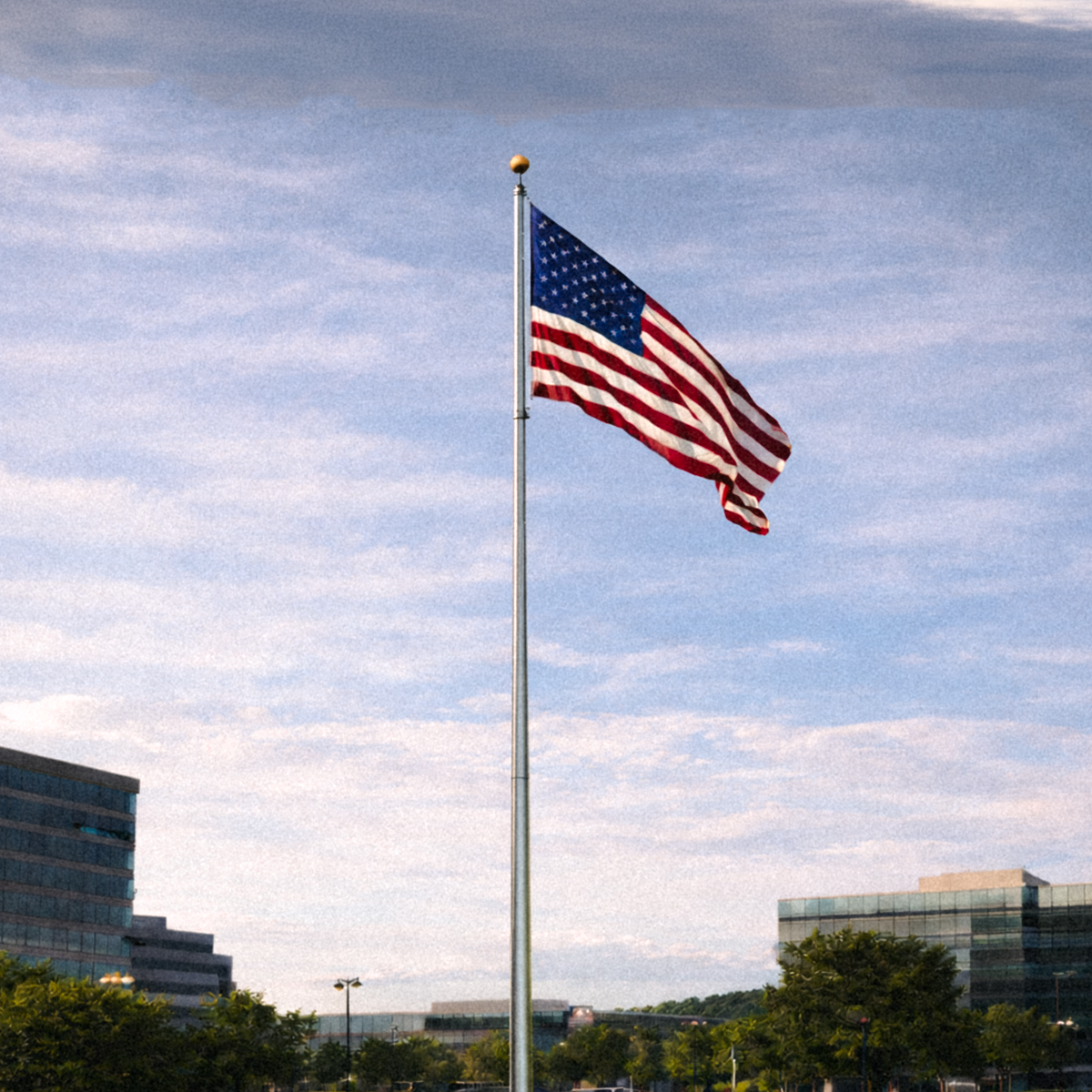 American flag waving on a flagpole against a blue sky with clouds, buildings, and trees in the background.