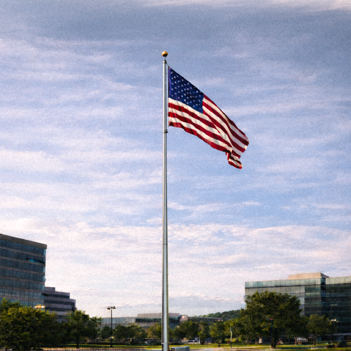 American flag waving on a flagpole against a blue sky with clouds, with buildings and trees in the background.