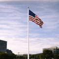 American flag waving on a flagpole against a blue sky with clouds, with buildings and trees in the background.