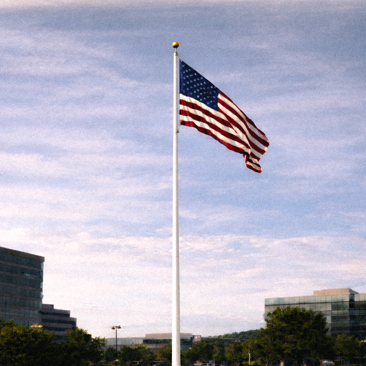 American flag waving on a flagpole against a blue sky with clouds, with buildings and trees in the background.