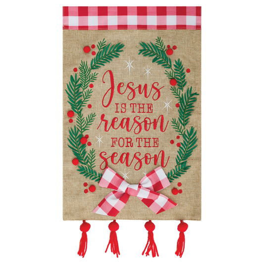 Decorative garden flag banner with 'Jesus is the reason for the season' text, surrounded by greenery and a bow on a burlap background.