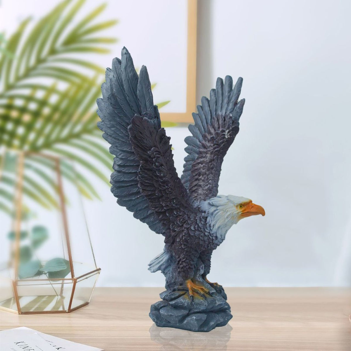 Eagle statue with open wings on a wooden surface with a blurred plant and frame in the background