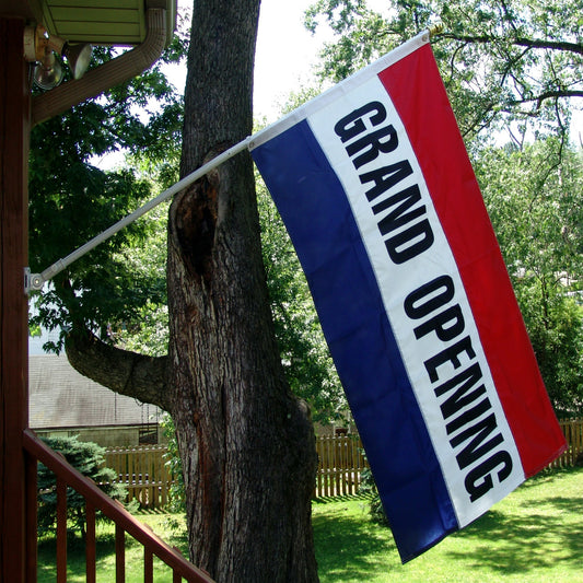 Grand Opening Flag - 3ft x 5ft - Secondary Image - United States Flag Store A 3ft x 5ft grand opening flag displayed on a flagpole, featuring red, white, and blue colors with bold black lettering, attached to a tree and a house in the background.